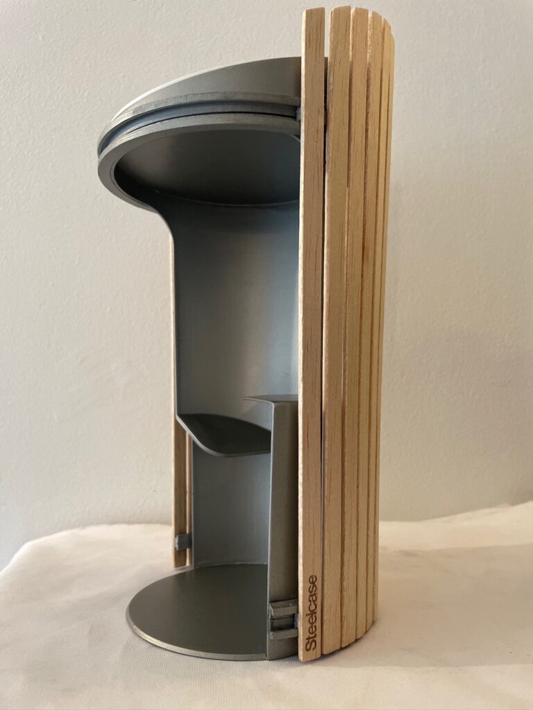 Prototype cylindrique en métal avec porte lamellée en bois partiellement ouverte, révélant un espace intérieur.